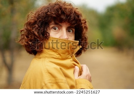 Covering face and making big eyes. Autumn portrait of young attractive fashionable happy curly girl at orange jacket looking straight in camera. European autumn fall park photo shoot