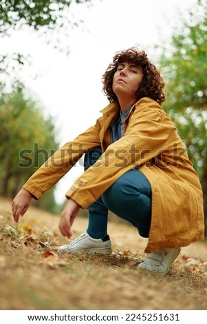 European autumn fall park photo shoot. Autumn portrait of young attractive fashionable sitting curly girl wearing orange jacket looking straight in camera. High quality vertical photo