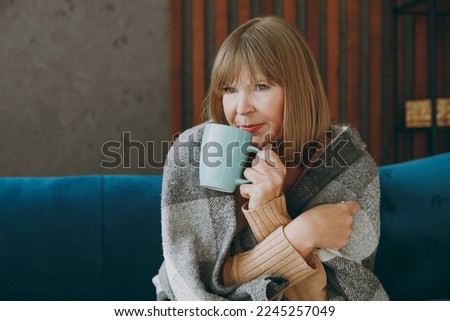 Elderly sad woman 50s years old wrap in plaid sits on sofa drink tea stay at home flat spending free spare time in living room indoor Healthy lifestyle ill sick disease treatment cold season concept Royalty-Free Stock Photo #2245257049