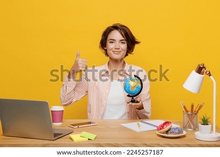 Young happy employee business woman wear casual shirt sit work at office desk with pc laptop hold Earth world globe show thumb up isolated on plain yellow color background. Achievement career concept