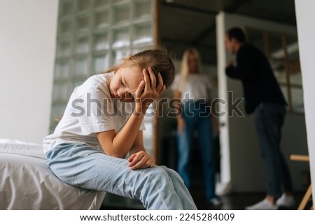 Front view of sad lonely little girl looking away, crying suffering sitting on sofa during parents quarrelling and fighting in living room on background. Concept of family problems, conflict, crisis. Royalty-Free Stock Photo #2245284009