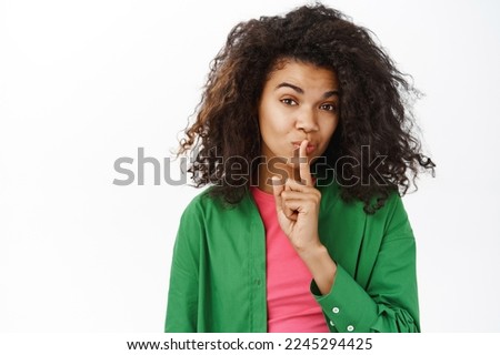 Shh keep it secret. Young teenage curly girl, makes hush, shush taboo sign, press one finger to lips and looks mysterious, white background