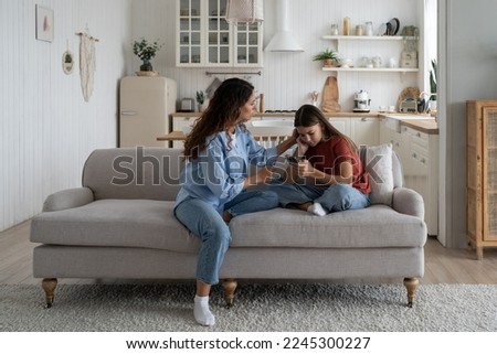 Loving mother sitting with sad daughter on sofa at home, helping child to cope with friendship breakup. Disappointed teen girl looking at phone screen, sharing feelings with mom. First love concept Royalty-Free Stock Photo #2245300227