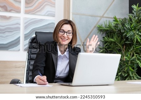 Portrait of successful and attractive business woman working on laptop in her office. Positive female employee in black suit smiling and sitting at the desk in her modern workplace