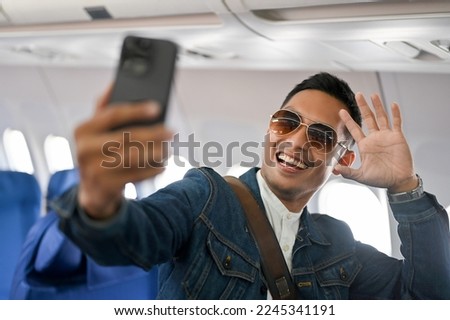 Handsome and joyful Asian male travel blogger or tourist wearing sunglasses taking selfie and recording video of himself by his smartphone during the flight. vacation, holiday, transportation