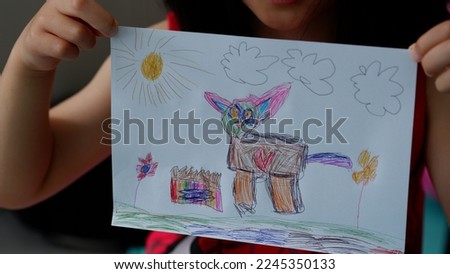 A litte girl shows the result of her drawing. Drawing a cat with a box of food, flowers, sun and clouds.