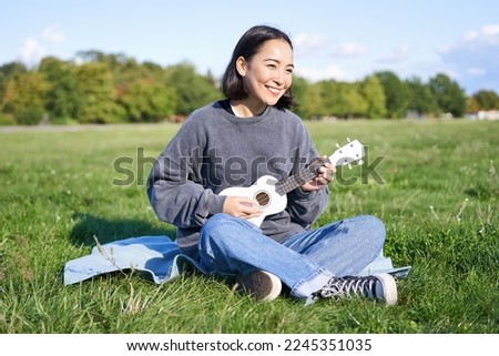 Beautiful asian girl sitting in park, playing ukulele and singing, relaxing outdoors on sunny spring day. Royalty-Free Stock Photo #2245351035