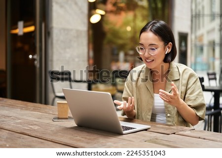 Young woman sitting on online meeting in outdoor cafe, talking to laptop camera, explaining something, drinking coffee. Digital nomad Royalty-Free Stock Photo #2245351043