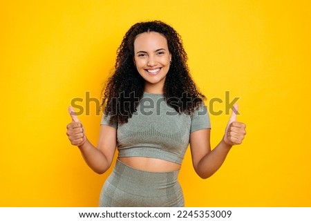 Photo of positive happy lovely curly haired brazilian or hispanic sporty woman in sportswear, showing thumb-up gesture, looks at camera, smiling, standing on isolated orange background
