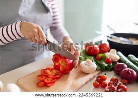 Cooking concept. Unrecognizable woman in grey apron making healthy dinner, cropped of lady preparing delicious meal at home, cutting vegetables on chopping board, kitchen interior, closeup