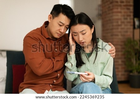Upset mature asian husband comforting his depressed young wife with negative pregnancy test, sitting on sofa at home. Korean couple cannot have baby, suffering from infertility indoors Royalty-Free Stock Photo #2245369235