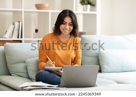 Smiling Young Arab Female Student Using Laptop At Home And Taking Notes, Happy Middle Eastern Woman Study With Computer While Sitting On Couch In Living Room, Enjoying Online Education, Free Space