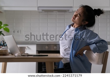 Unhappy Asian student girl suffering from lower back pain while studying on laptop from home, tired unhealthy female freelancer feeling pain or discomfort when sitting in poor posture long hours Royalty-Free Stock Photo #2245371579
