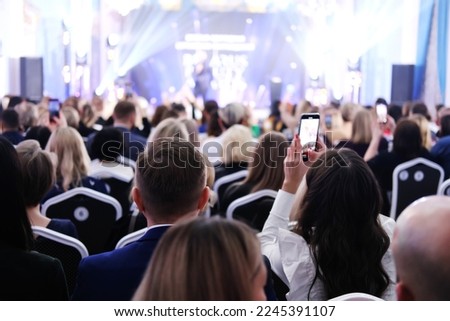 Guests in evening attire sit in a spotlighted lobby and look at the stage. Girl filming an event on the phone. Horizontal photo