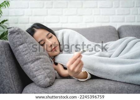 Winter Illnesses. Young sick Asian woman taking temperature with thermometer while lying under blanket on sofa at home. 