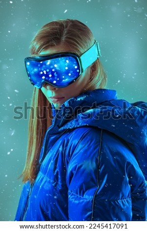 Portrait of a beautiful girl in a blue down jacket and ski goggles under a snowfall. Skiing sport. Winter fashion.