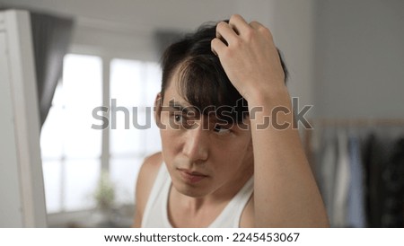 unhappy taiwanese man checking hairstyle in the mirror is shocked and worried about hair loss problem while preparing for a date in the bedroom at home. Royalty-Free Stock Photo #2245453067