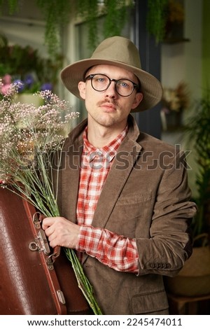 Attractive male poet or writer in eyeglasses and vintage clothes staying with flower bouquet and looking at camera in flower store. High quality vertical image