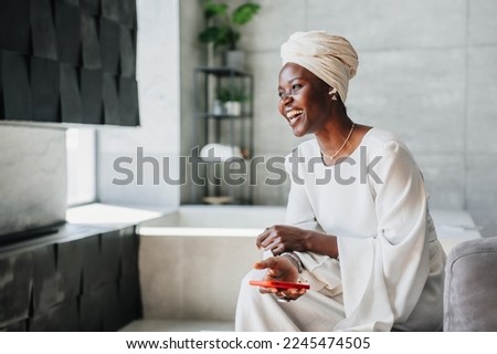 Cheerful African woman in white dress and turban holds phone sitting at home on couch. Happy African American businesswoman laughs looks away being satisfied. People candid emotions. Woman in hotel. Royalty-Free Stock Photo #2245474505
