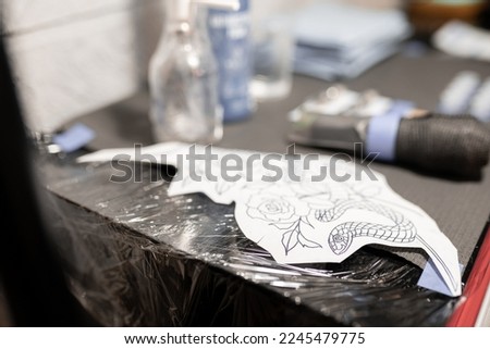 A tattoo design stencil is resting on the work table next to the other tools before tattooing Royalty-Free Stock Photo #2245479775