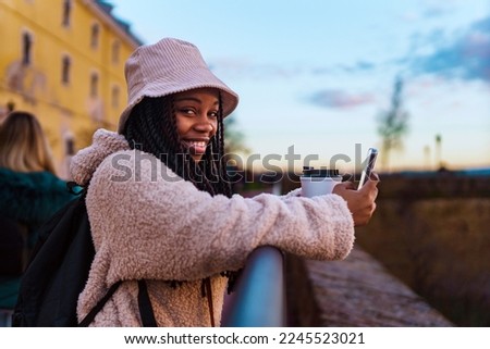 Portrait of a beautiful black woman smiling excitedly at the camera, she's wearing a bucket hat and a backpack, she's drinking coffee and taking pictures with her phone Royalty-Free Stock Photo #2245523021