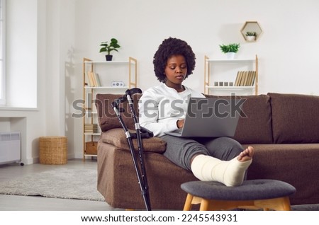 Attractive African American woman with broken leg in cast is sitting on the sofa in the living room with laptop in hands. Recovery and rest at home after rehabilitation of broken leg. Crutches nearby. Royalty-Free Stock Photo #2245543931