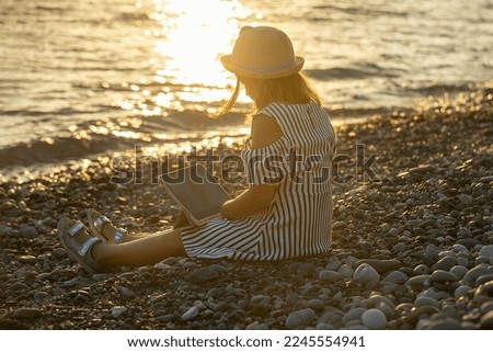 girl schoolgirl tourist sits on the seashore in the evening, at sunset and looks at the tablet, she is dressed in a blue striped dress and a straw hat
