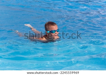 Boy child in swimming goggles with float foam board tool swim in pool in sunny day. Safe pool training, summer holiday, family vacation, travel, water sport concept Royalty-Free Stock Photo #2245559969