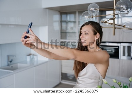 Adorable caucasian woman in nightie makes selfie at kitchen toothy smiles looks at screen, makes video call at home. Pretty Italian female model holds cell phone. Domestic life, weekend leisure.