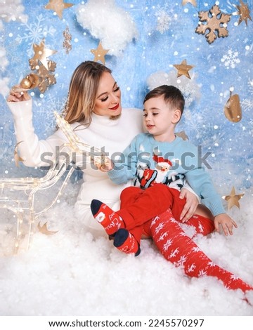 Little boy in sweater with pregnant mom in the winter forest. Winter wonderland setup with snow, stars and Christmas deer props. 