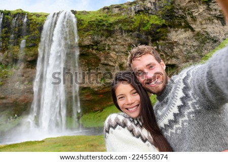 Iceland couple selfie wearing Icelandic sweaters by Seljalandsfoss waterfall on Ring Road in beautiful nature landscape on Iceland. Woman and man model in typical Icelandic sweater. Multiracial couple