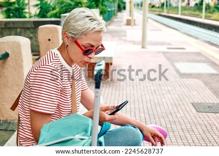 Blonde woman tourist using smartphone for social media check-in, city transport map or paying online ticket service while waiting for train on railway station.Modern travel app technology.
