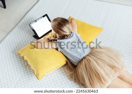 Cute little caucasian girl with blonde hair in fashionable clothes sitting at home during coronavirus pandemic quarantine and using tablet. 