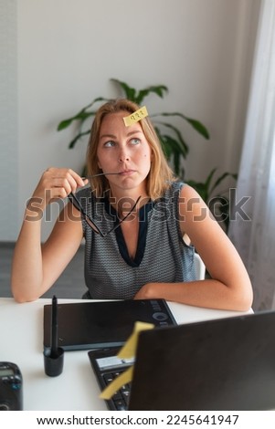 A young woman sitting at her Desk, holding glasses and thinking about something. On the forehead sticker with question marks. View from the top.