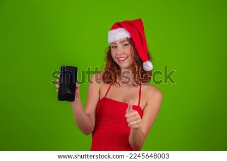 Happy young woman in red Santa Claus hat holds mobile phone in hand and makes a thumbs up sign