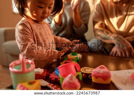 Focused asian little girl playing with toy figures at table near blurred cropped mother and father on sofa at home. Domestic entertainment and leisure. Modern childhood lifestyle. Sunny day