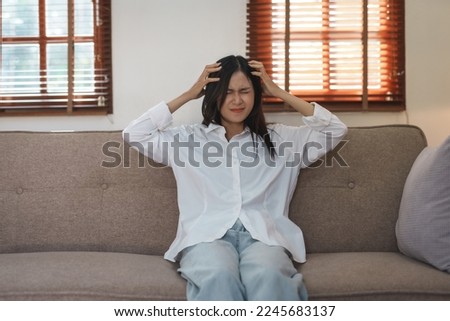 Young Asian woman suffering from stress from an existing disease having a headache. Migraine sitting alone on the sofa at home frustrated. Desperate Asian woman feeling lonely, tired. Royalty-Free Stock Photo #2245683137