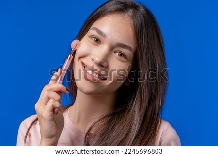 Teen girl doing face massage with rose quartz stone roller on blue background. Facial self care, beauty rituals, cosmetology, anti aging and anti-wrinkle treatment. High quality photo