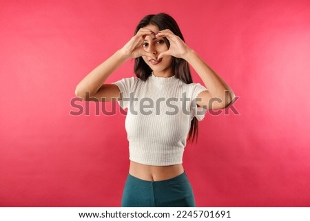 Cute brunette woman isolated over red background doing heart shape with hand and fingers looking through sign. Smiles with love. Makes a heart sign with hands and looks at the camera, at you.