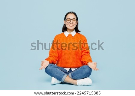 Full body fun young woman of Asian ethnicity wear orange sweater glasses hold spreading hands in yoga om aum gesture relax meditate try to calm down isolated on plain pastel light blue cyan background