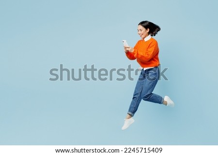 Full body young woman of Asian ethnicity wear orange sweater glasses jump high hold in hand use mobile cell phone isolated on plain pastel light blue cyan background studio. People lifestyle concept Royalty-Free Stock Photo #2245715409