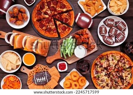 Super Bowl or football theme food table scene. Pizza, hamburgers, wings, snacks and sides. Overhead view on a dark wood background. Royalty-Free Stock Photo #2245717599
