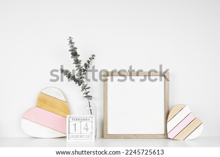 Mock up square wood frame with rustic Valentines Day wooden heart decor, eucalyptus branch and calendar. White shelf against a white wall. Copy space.