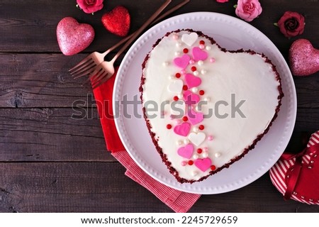 Valentines Day theme red velvet heart shaped cake with white icing and colorful heart decoration. Top view over a dark wood background.