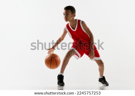 Boy in red uniform training, playing basketball, dribbling over grey studio background. Motivated sportsman. Concept of energy, professional sport, motion, action, hobby, competition, achievement.