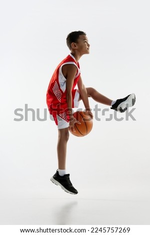 Portrait of boy in red uniform training, playing basketball over grey studio background. Jumping with ball. Concept of energy, professional sport, motion, action, hobby, competition, achievement.