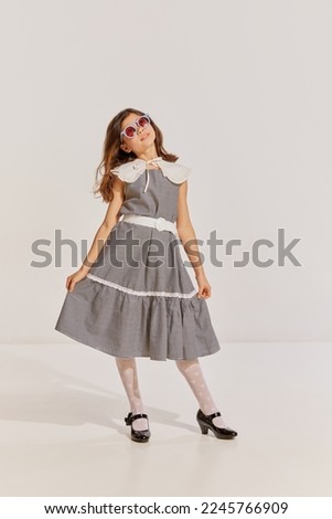 Portrait of beautiful teen girl in vintage dress and funny sunglasses posing over grey background. Concept of childhood, friendship, fun, lifestyle, fashion, retro style, emotions