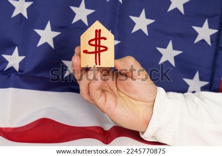 Business concept. Against the background of the USA flag, a man's hand holds a wooden house with the image of the dollar symbol.