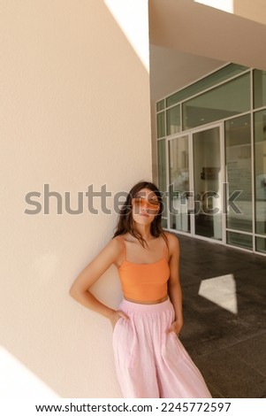 Vertical image with calm young caucasian woman posing looking at camera with hands in pockets. Model with brunette hair wears top, pants and sunglasses. Relaxing concept.