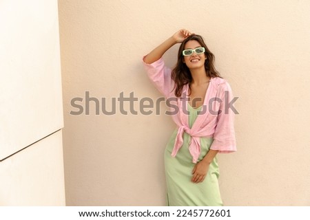 Positive young caucasian woman smiling with teeth posing at camera near building wall. Model wears shirt, sundress and sunglasses. Concept of stylish vacation. Royalty-Free Stock Photo #2245772601